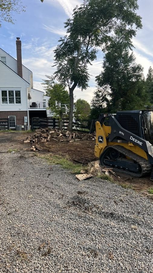 John Deere 317G next to a stacked woodpile after tree removal